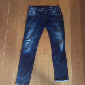 Replay Anbass jeans blå, stl 32/32 - Snygga Replay Anbass jeans i mörkblå tvätt med feta slitningar och lätt trasiga detaljer vid knäna. Klassisk modell med smal passform och stretchig känsla. Perfekta för dig som gillar en modern och avslappnad stil.