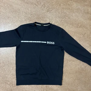 Svart BOSS sweatshirt med tryck - Svart sweatshirt från BOSS i storlek M. Tröjan har ett vitt tryck med texten 'BOSS' och en horisontell linje över bröstet. Klassisk rund halsringning och långa ärmar. Perfekt för en clean och stilren look. Har en vit fläck på ena ärmen som inte går bort. 