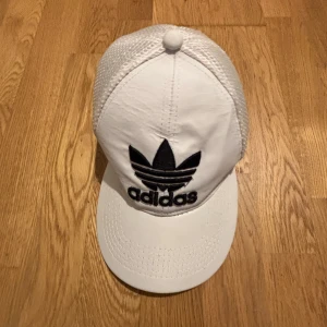 Vit Adidas keps med mesh - Snygg vit keps från Adidas med svart broderad logga framtill. Kepsen har klassisk böjd skärm och mesh-paneler på sidorna och bak för extra ventilation. Perfekt för dig som gillar sportig och clean stil.