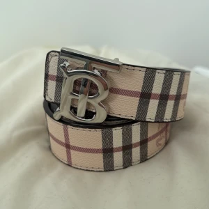   Burberry bälte - Säljer ett klassiskt bälte från Burberry med deras ikoniska rutiga mönster i beige, svart och rött. Bältet har ett stort silverfärgat B-spänne och är tillverkat i skinn. Perfekt accessoar för att lyfta din outfit. Den är 110cm,helt ny och ej använd kom dm för mer.