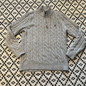 Grå kabelstickad tröja från Polo Ralph Lauren - Säljer en grå kabelstickad tröja från Polo Ralph Lauren med halv dragkedja och hög krage. Tröjan har långa ärmar, ribbade muddar och en broderad logga på bröstet. Perfekt för kyligare dagar.