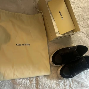 Svarta sneakers från Axel Arigato - Säljer ett par svarta sneakers från Axel Arigato med vit sula och diskret logga på sidan. Skorna har snörning och är tillverkade i skinn. Kommer med originalpåse och låda.
