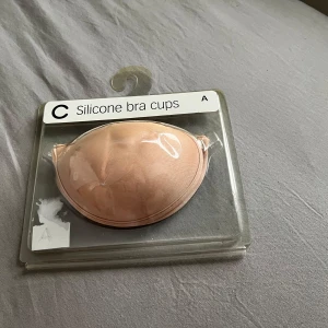 Beige silikon bh-cups 75a - Säljer ett par beige silikon bh-cups som är självhäftande och axelbandslösa. Perfekt när du vill ha en osynlig look under kläderna. De har en smidig knäppning framtill och passar storlek A.