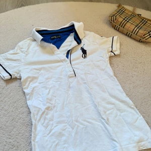 Vit pikétröja från Ralph Lauren - Vit pikétröja från Ralph Lauren med klassisk krage och knappar framtill. Tröjan har blå detaljer vid krage och ärmslut samt den ikoniska loggan broderad på bröstet. Perfekt för en sportig och stilren look.