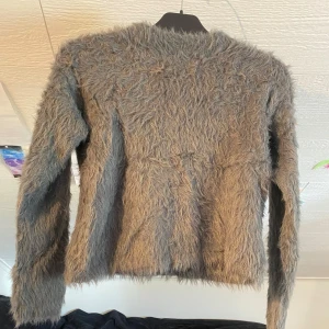 Grå fluffig tröja med lång lugg - Säljer en unik grå tröja med lång, lurvig pälslook. Tröjan har rund halsringning och långa ärmar. Perfekt för dig som vill sticka ut och ha något riktigt annorlunda i garderoben!