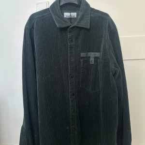 MörkGrön corduroy manchester-skjorta från Stone Island - Säljer en mörkgrön manchester-skjorta från Stone Island med klassisk krage och knappstängning framtill. Skjortan har en bröstficka med logotypdetalj och knäppning. Perfekt för dig som gillar streetwear och vill ha något unikt.