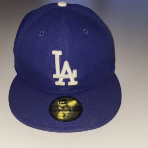 Blå LA Dodgers keps från New Era  - Säljer en blå LA Dodgers keps från New Era  med vit broderad LA-logga framtill och MLB-logga bak. Klassisk flat peak och broderade ventilationshål. Perfekt för dig som gillar sportig stil. Det är en fitted cap och är riktig snygg för dig som vill ha en lite mer street stil.