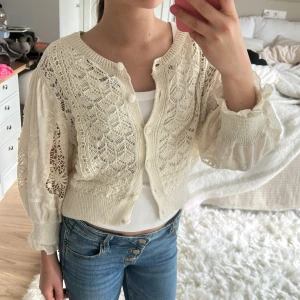 Vit spetskofta med knappar - Säljer en söt vit kofta med spetsdetaljer och knappar framtill. Koftan är croppad och har långärmade, lätt puffiga ärmar med ribbade muddar. Perfekt att slänga över ett linne eller t-shirt för en extra touch.