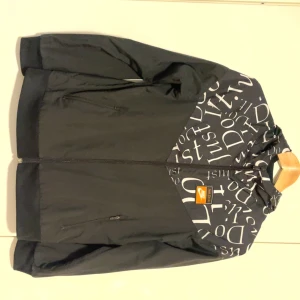 Svart och vit vindjacka från Nike - Nike vindjacka i svart med vit textprint på axel och huva. Jackan har dragkedja framtill och en orange Nike-logga med 'Just Do It.' på bröstet. Perfekt för en sportig och stilren look.