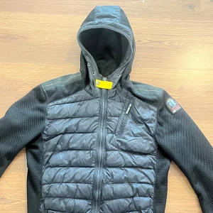 Svart hybridjacka med huva från Parajumpers - Säljer en svart Parajumpers hybridjacka med quiltad front och ärmar i stretchigt material. Jackan har huva, dragkedja framtill och en bröstficka med dragkedja. Märkespatch på ena ärmen och gul detalj vid halsen. Litet hål vid baksidan men inget man tänker på