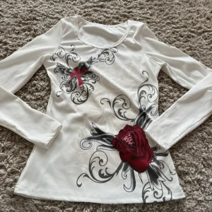 Vit långärmad topp med tryck av ros och kors - Säljer en vit långärmad topp med stora tryck av en röd ros och ett rött kors med gråa och svarta detaljer. Tröjan har rund halsringning och är figurnära i modellen. Perfekt för dig som gillar unika prints!