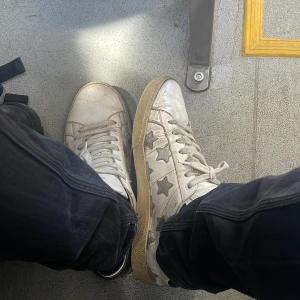 Vita sneakers med stjärnor från Saint Laurent, 700 så får ni dem vid snabb affär!! - Säljer ett par vita sneakers från Saint Laurent med grå stjärnor på sidorna och beige sula. Skorna har snörning och rund tå. Märket syns på plösen och insidan. Perfekta för dig som gillar en unik och trendig look. De är i acceptabelt skick, lite defekter, går lätt att tvätta så att de blir riktigt snygga. Det svarta är bara en extra sula jag använder. Fråga bara på!