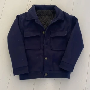 Mörkblå Fältjacka - Mörkblå jacka med krage och stora fickor framtill. Jackan har knäppning med knappar och quiltat innerfoder. Perfekt för kyligare dagar och har en klassisk, stilren look. För 12-13 år.