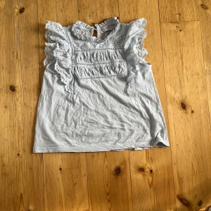 Ljusblå blus med volang från Lindex - Säljer en superfin ljusblå blus från Lindex. Blusen har volangdetaljer på axlarna samt broderade spetsdetaljer framtill. Perfekt för vår och sommar! Aldrig använd.
