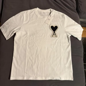 Vit t-shirt med hjärtdetalj från Ami - Säljer en äkta vit t-shirt från Ami med en stor svart hjärtdetalj och bokstaven A på bröstet. T-shirten har normal passform och är tillverkad i mjuk bomull. Perfekt för en stilren och trendig look. Lite stor i storleken så passar även för dig som har storlek S.