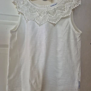 Vit ärmlös topp med spetskrage från Polarn O. Pyret - Säljer en söt vit ärmlös topp från Polarn O. Pyret med dekorativ spetskrage. Toppen har rund halsringning och knäppning bak i nacken. Perfekt för en stilren och klassisk look. Ny aldrig använd.kommer med en extra knapp på tätvättrådet