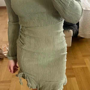 Ljusgrön långärmad klänning med volang - Säljer en ljusgrön långärmad klänning med rynkade detaljer och volangkant nertill. Klänningen har en tight passform och är tillverkad i ett plisserat material. Snygg knytning på sidan ger extra detalj.