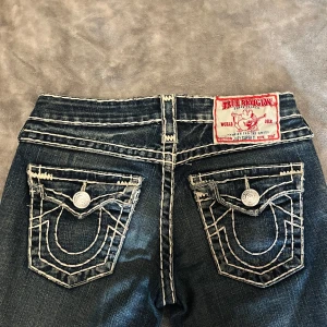 True Religion jeans med kontrastsömmar - Säljer ett par klassiska True Religion jeans med tydliga vita kontrastsömmar och ikoniska fickdetaljer bak. Jeansen har bootcut-modell och är tillverkade i blå denim. Märkespatch bak i midjan och knappar med logga. Jätte fina älskar dem men säljer på grund av att dem har blivit lite för stora för mig 💞pris kan diskuteras 