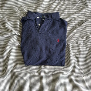 Mörkblå pikétröja från Ralph Lauren - Säljer en klassisk mörkblå pikétröja från Ralph Lauren med röd broderad logga på bröstet. Tröjan har korta ärmar och en stilren krage med knappar. Perfekt för en avslappnad men stilren look.