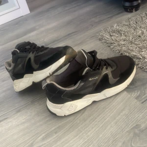 Svarta sneakers från Gant - Säljer ett par svarta sneakers från Gant med vita, chunky sulor och svarta snören. Skorna har detaljer i grått och vitt samt en sportig design. Perfekta för en avslappnad och trendig stil.