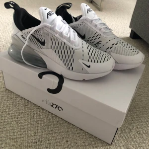 Nike Air Max 270 White - Nike Air Max 270 i vitt med svarta detaljer och stor synlig luftbubbla i hälen. Ovandelen är i mesh med svarta loggor och detaljer, samt vit sula. Snygga och sportiga sneakers med modern design.