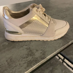 Beige sneakers från Michael Kors med gulddetaljer - Snygga beige sneakers från Michael Kors med guldfärgade detaljer och meshpanel på sidan. Skorna har snörning och en vit platt sula. Perfekta för en trendig och sportig look.