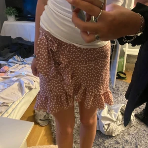 Rosa omlottkjol med vita prickar och volang - Supersöt rosa omlottkjol med vita blommor och volangkant. Kjolen har knyt i midjan och är kort/medel i modellen. Perfekt till sommarn 💕💕