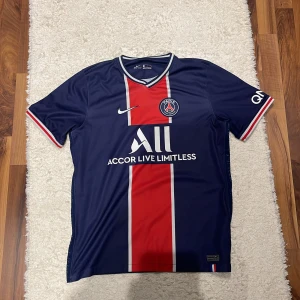 PSG hemmatröja 2020/21 - Paris Saint-Germain fotbollströja i mörkblått med röd och vit rand i mitten. Nike-logga och PSG-märke på bröstet, samt sponsortryck. Kortärmad modell med V-ringning och detaljer i vitt och rött vid ärmslut och krage.