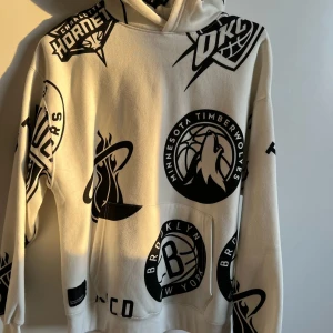 Vit hoodie med NBA-lagloggor - Säljer en vit hoodie från Nike med stora svarta tryck av olika NBA-lagloggor, bland annat Miami Heat, Brooklyn Nets och Minnesota Timberwolves. Tröjan har huva och känguruficka framtill. Perfekt för basketfans!