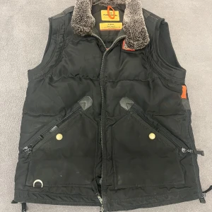 Svart dunväst från Parajumpers - Säljer en svart dunväst från Parajumpers med dragkedja och två stora snedställda fickor framtill. Västen har en mjuk, grå fuskpäls vid kragen och orange detaljer. Perfekt för lager-på-lager under kyliga dagar.