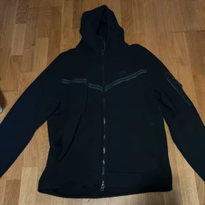 Svart Nike tech  - Säljer en svart  Nike tech i storlek L. Jackan har hel dragkedja framtill, huva och diskreta reflexdetaljer över bröstet. Perfekt för dig som gillar sportig och enkel stil.