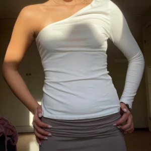 Vit oneshoulder topp  - Säljer en vit, långärmad oneshoulder topp i stretchigt material från Nelly. Toppen har en axel bar och en lång ärm, vilket ger en snygg och modern look. Perfekt att styla med kjol eller jeans för en trendig outfit. Mycket bra skick!