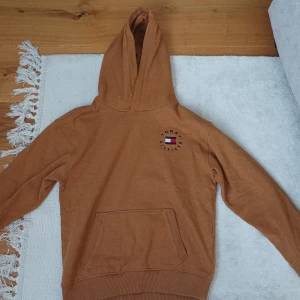 Brun hoodie från Tommy Hilfiger - Säljer en brun hoodie från Tommy Hilfiger med klassisk logga på bröstet. Tröjan har huva och en stor magficka framtill. Perfekt för en avslappnad och stilren look. OANVÄND!