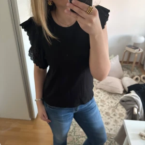 Svart topp med volangärmar från Zara - Svart topp från Zara med korta volangärmar i spets. Toppen har rund halsringning och är tillverkad i ett mjukt material. Perfekt för dig som vill ha en enkel men ändå detaljrik topp. Storlek M men passar också S