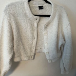 Vit fluffig cardigan kofta från Gina Tricot - Mysig vit cardigan kofta med fluffig känsla och stora knappar framtill. Croppad modell med lång ärm och rund halsringning. Perfekt för lager-på-lager och enkel att styla till olika outfits.