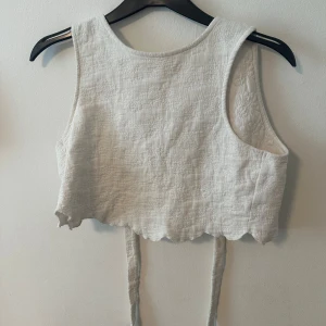Vit croppad linnetopp med knyt - Säljer en söt croppad väst i vit linne med knyt baktill. Västen har en vågig nederkant och är ärmlös, perfekt för varmare dagar. Enkel och stilren design som passar till många outfits.