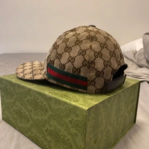 Beige keps från Gucci med monogram - Säljer en beige keps från Gucci med klassiskt GG-monogram över hela kepsen. Kepsen har gröna och röda ränder på sidan och justerbar rem baktill i svart läder. Perfekt accessoar för en trendig look.