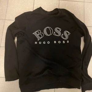 Svart sweatshirt från Hugo Boss - Säljer en svart sweatshirt från Hugo Boss med stor vit logga på bröstet. Tröjan har rund halsringning och långa ärmar. Perfekt för en stilren och avslappnad look.