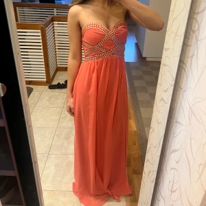 Balklänning i Korallfärg - Säljer en korallfärgad lång balklänning från Cotton Club med axelbandslös design och glittriga stenar över bysten. Klänningen har markerad midja och ett flytande, elegant fall. Perfekt för bal eller festliga tillfällen. Jag som bär den är vanligtvis en storlek s så skulle gissa på att den är kring den storleken! Går även att lägga till band om man känner för det för det finns möjlighet på det i insidan av klänningen men har inga band! Man får även med den matchande sjalen på andra bilden!!