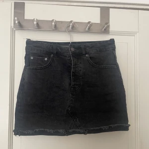 Svart jeanskjol med rå kant - Säljer en svart jeanskjol från perfect jeans med fem fickor och rå, fransad kant nedtill. Kjolen har klassisk knapp- och dragkedjestängning framtill och är i en kort modell. Perfekt till vardags med en t-shirt eller hoodie.
