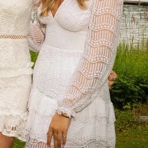 Vit studentklänning från NA-KD - Superfin vit student klänning från NA-KD med v-ringning och långa ärmar. Klänningen har spetsdetaljer, volanger och ett romantiskt mönster. Perfekt för sommarens tillfällen och studentfirande.