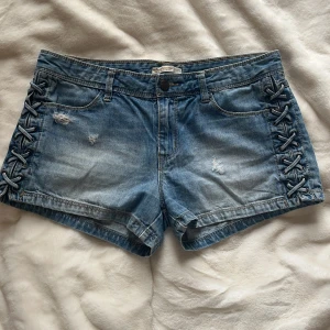 Blå jeansshorts med snörning i sidorna - Säljer ett par ljusblå jeansshorts med snygga snörningsdetaljer längs båda sidorna. Shortsen har fem fickor, slitna detaljer framtill och klassisk jeanslook. Perfekta för en vacker sommardag!!🥰🥰 