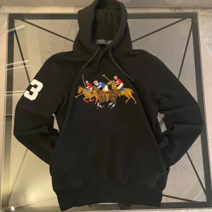 Svart hoodie från Polo Ralph Lauren med broderat polomotiv - Säljer en svart hoodie från Polo Ralph Lauren med stort broderat polomotiv på bröstet och siffran 3 på ärmen. Tröjan har huva med dragsko och ribbade muddar. Perfekt för dig som gillar sportig stil och klassiska märken.