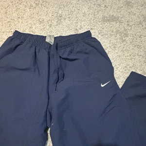 Vintage Marinblå träningsbyxor från Nike - Säljer ett par marinblå träningsbyxor från Nike med resår i midjan och snörning. Byxorna har dragkedja vid bensluten och en liten vit Nike-logga på ena benet. Perfekta för sport eller chill.