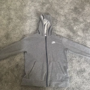 Grå hoodie från Nike med dragkedja - Säljer en grå hoodie från Nike i storlek 147-158. Tröjan har dragkedja framtill, huva och två fickor. Klassisk Nike-logga på bröstet och mjukt material på insidan. Perfekt för en avslappnad stil. Sälj pågrund av att den inte passar längre
