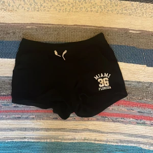 Svarta shorts med Miami-tryck - Svarta shorts med vit snörning i midjan och tryck på ena benet där det står 'Miami 36 Florida'. Mjukt material, perfekt för chill dagar eller träning. Sportig vibe och kort modell som sitter skönt på kroppen.