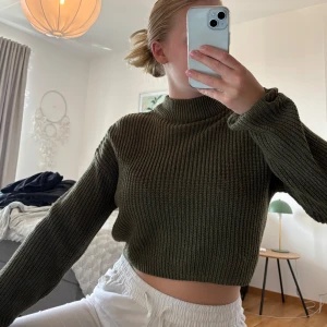 Olivgrön croppad stickad tröja - Säljer en olivgrön croppad stickad tröja med hög hals och långa ärmar. Tröjan har en ribbad struktur, enkel och stilren design som passar till mycket.