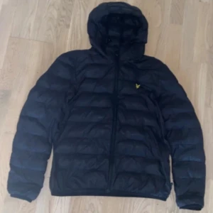 Lyle & Scott Jacka - Säljer en svart pufferjacka från Lyle & Scott med huva och dragkedja. Jackan har ett diskret logomärke på bröstet och är vadderad för extra värme. Perfekt för kyliga dagar och enkel att matcha med olika outfits. Jackan har ett hål på fickan som inte syns när man har på sig den och den har ett hål på baksidan av jackan som kan synas lite mer.