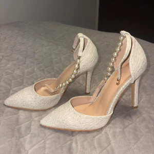Vita pumps med pärldetalj och spetsig tå - Säljer ett par eleganta vita pumps (glitter) med hög klack och spetsig tå. Skorna har en dekorativ rem med pärlor runt vristen och en fin struktur i materialet. Perfekta för dig som vill sticka ut med stilrena detaljer.Kan även passa storlek 37
