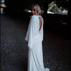 Vit glittrig långklänning med öppen rygg - Säljer en vit långklänning med glittrigt material och djup v-ringning. Klänningen har vida ärmar och en elegant öppen rygg. Perfekt för dig som vill sticka ut med en glamorös look.